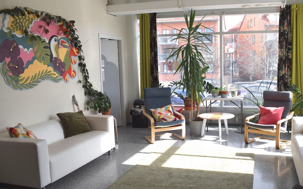 A room with a sofa, two armchairs, houseplants, and a community artwork on the wall.