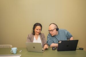 Kaksi henkilöä istumassa pöydän ääressä. Edessä kannettavat tietokoneet.