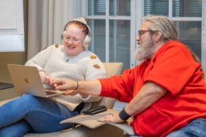 Kaksi hymyilevää henkilöä. Toisella on kannettava tietokone sylissä. Toinen osoittaa näytölle. 