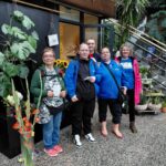 Viisi hymyilevää ihmistä seisomassa. Ympärillä viherkasveja.